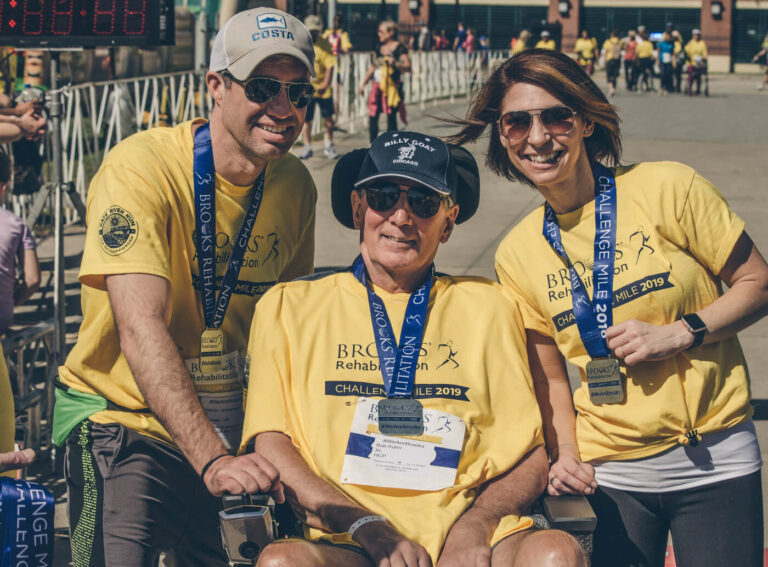 two men and a woman showing off their race medals after the Challenge Mile. event. The older participant in the middle is in a wheelchair.